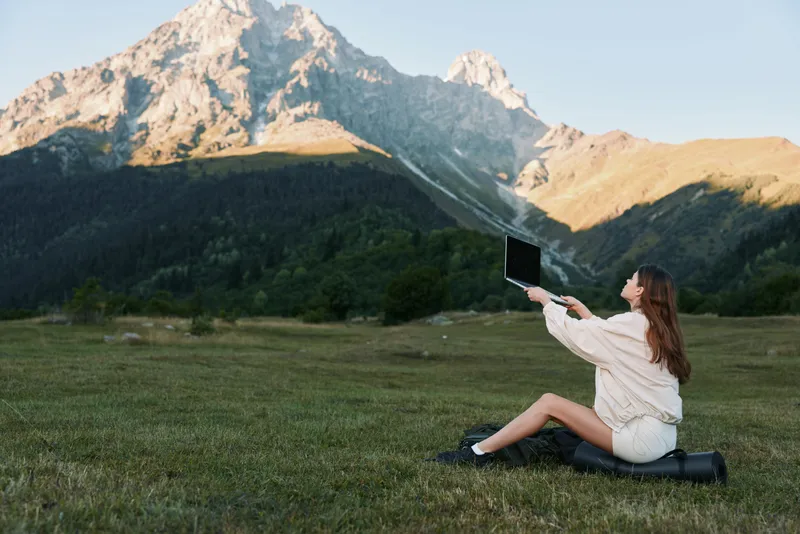 A woman sits on a small bag in a grassy field, using a laptop with a mountain backdrop. She appears focused and creative, conveying a productive outdoor work scene.