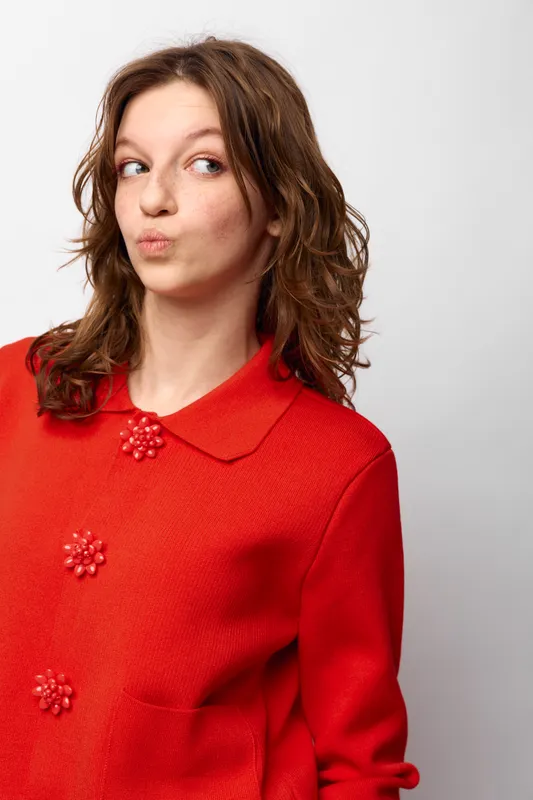 Young woman with curly brown hair wearing a red coat looks sideways with pursed lips. Studio portrait on white background. Concept of curiosity, doubt, and casual fashion.