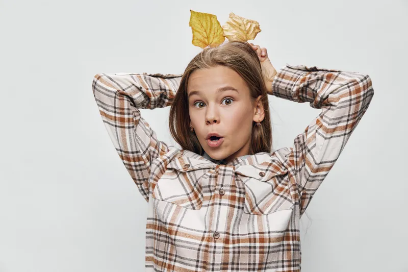 Girl wearing plaid shirt holding yellow leaves on head with surprised expression isolated on white background, autumn concept, casual kid with long hair and bright eyes.