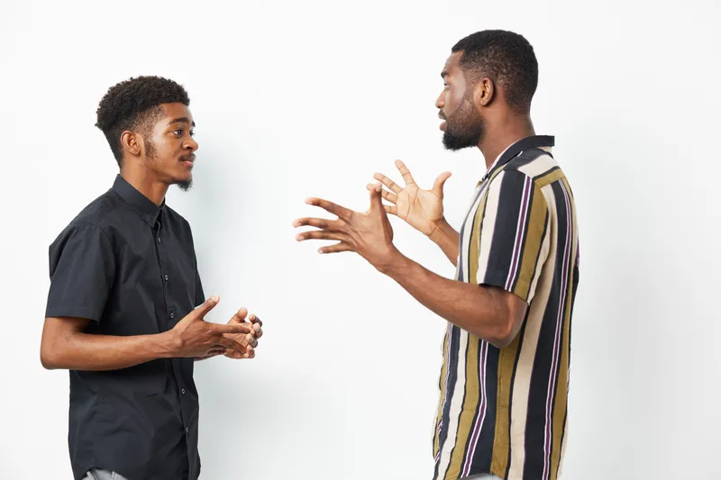 Two black men engaging in an animated conversation, showcasing expressions of interest and enthusiasm, wearing casual attire in a bright setting.