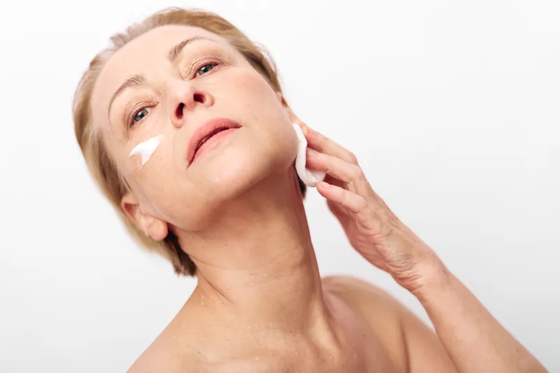 Mature woman with light skin applies white cream on cheek using cotton pad. Studio shot on white background. Concept of skincare, antiaging routine, and self-care for older adults.