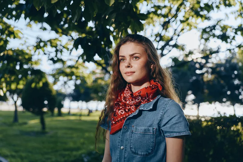 young woman with red bandana standing in green park wearing denim dress during sunny day with natural light and trees background