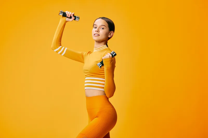 Woman in orange fitness outfit lifting small dumbbells in a vibrant studio, showcasing strength, energy and athletic training for sport and wellness.