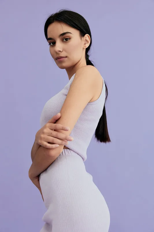 Young woman with dark hair wearing a sleeveless lavender dress stands confidently against a pastel purple background, showcasing minimalistic style and natural beauty.
