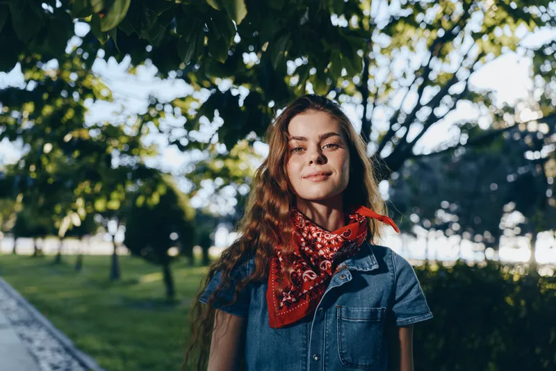 young woman with long hair wearing denim and red bandana outdoors in sunny park with trees and green grass, natural light, summer mood