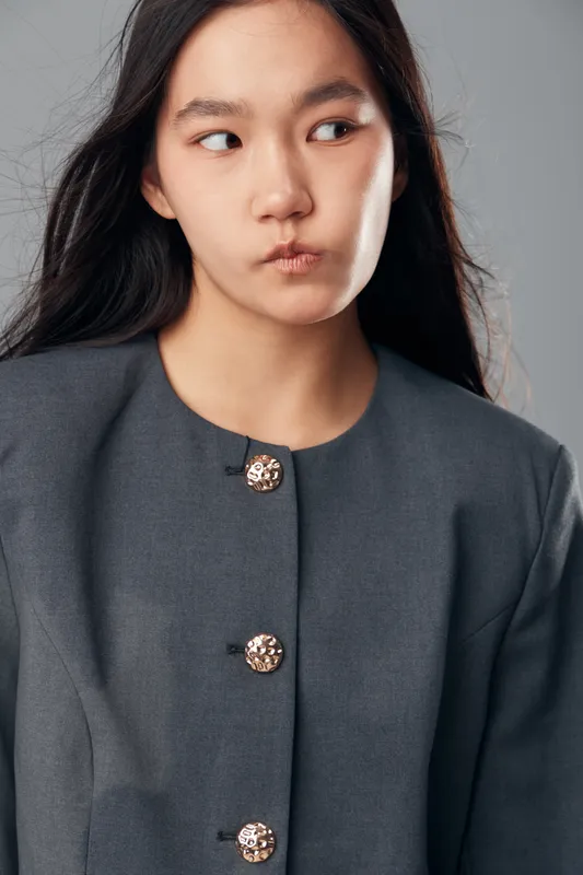 Portrait of young woman with thoughtful expression looking sideways, wearing gray jacket with decorative buttons on light gray background in studio. Asian female model with natural makeup.
