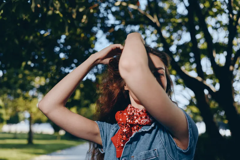 lifestyle woman in modern boho-western denim street style with red bandana stands outdoors under sunny trees in warm film color tone