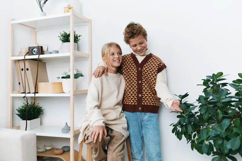 Two cheerful children wear cozy knitwear indoors, hugging near a shelf of plants and decor, exuding warmth, comfort, and playful sibling bond in a bright home setting.