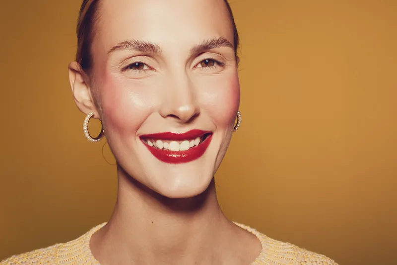 Smiling woman with red lipstick and hoop earrings poses against warm yellow background. Close-up portrait of confident female model with natural skin and bright makeup. Beauty concept.