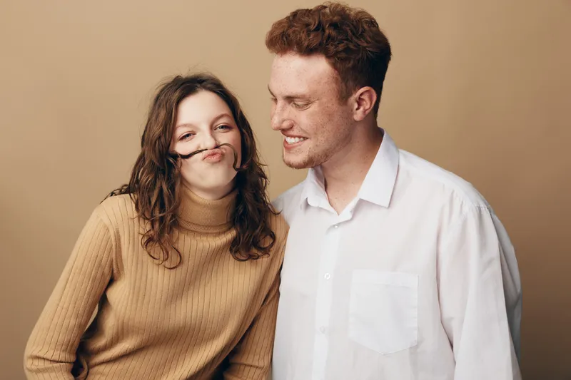 Young couple having fun together, woman making a mustache with her hair while man smiles. Casual clothing and beige background create a warm, playful mood of friendship and joy.