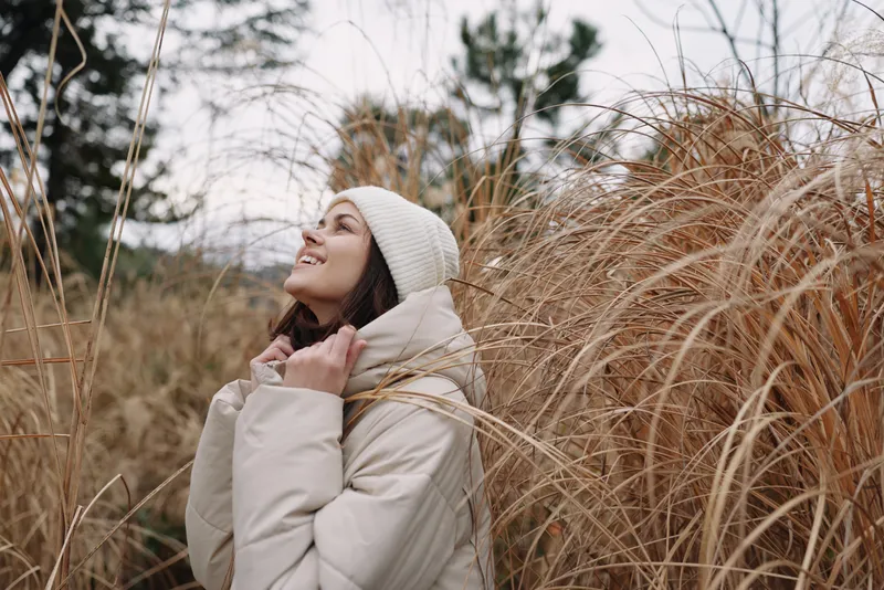 Woman outdoor autumn winter fashion coat scarf standing among tall dried grasses enjoying a chilly breeze in a peaceful natural moment