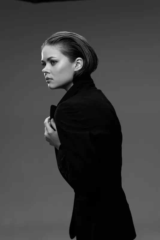 Black and white portrait of a serious woman wearing a coat in a studio setting with dramatic lighting.