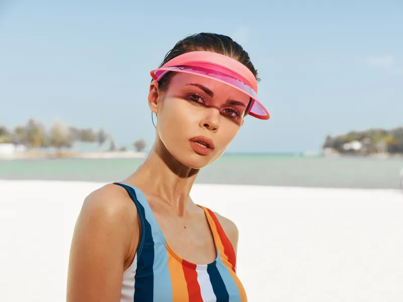 A stylish woman poses on a sunny beach wearing a colorful striped swimsuit and a pink visor, exuding confidence, summer chic, and bright, warm atmosphere.