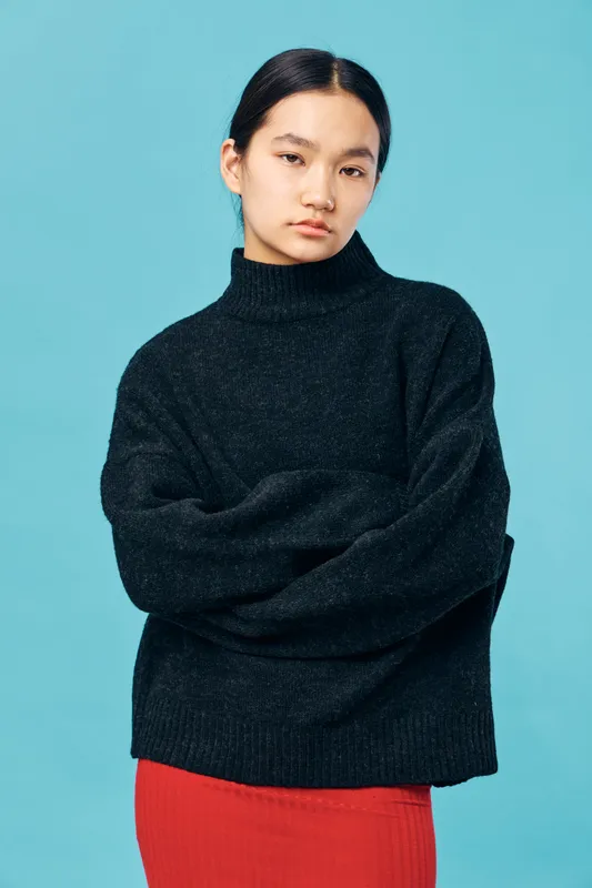 Woman with serious expression wearing black sweater and red skirt standing against blue background, posing with crossed arms in studio for fashion portrait or lifestyle.
