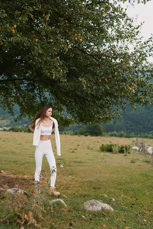 Woman outdoors in a white outfit stands in a grassy field beneath a large tree, capturing a casual lifestyle moment set in nature and open countryside.