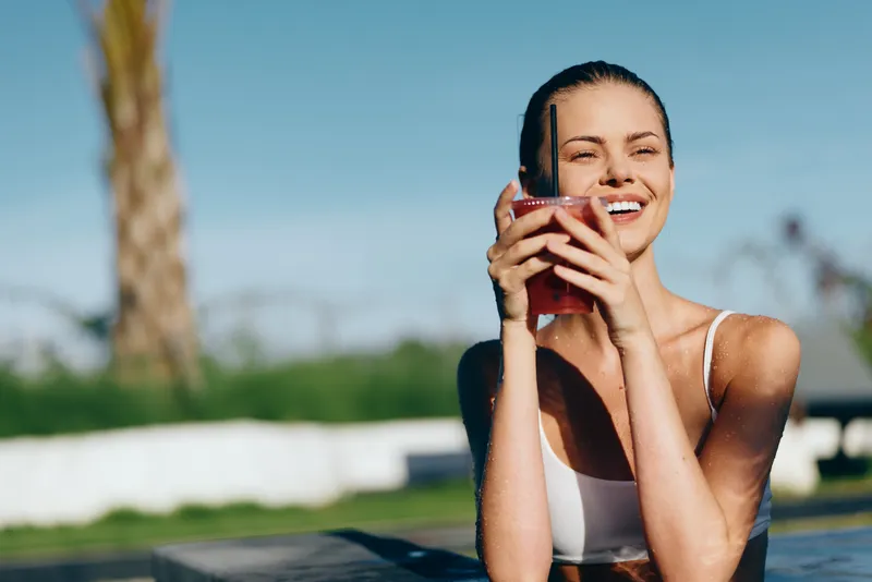 Young woman in white swimsuit enjoys fresh juice by the pool on a sunny day smiling with relaxed lifestyle and healthy outdoor activity background with palm tree and clear sky.