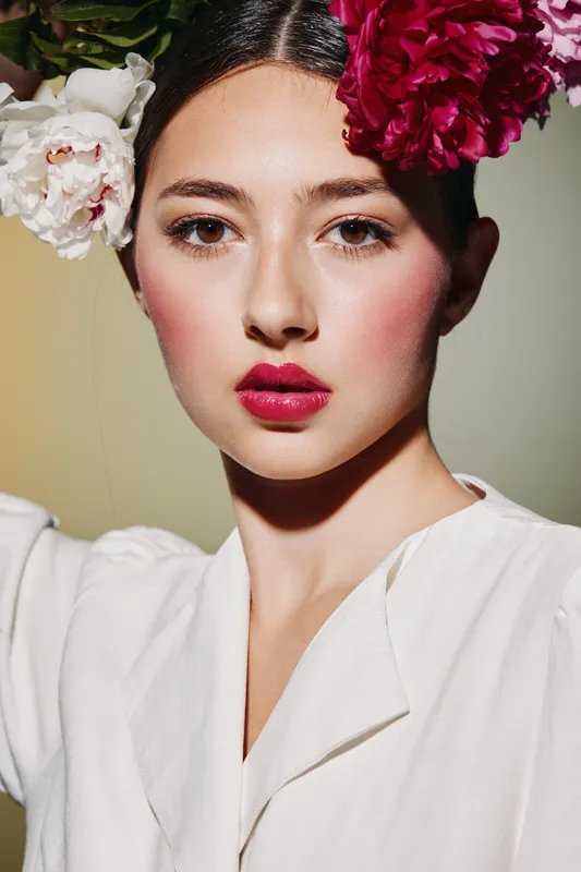 Portrait of a stylish woman with natural skin makeup and vibrant flowers in her hair. Shine highlights emphasis on her pink lipstick and fresh look. Beauty concept with fashion flair.