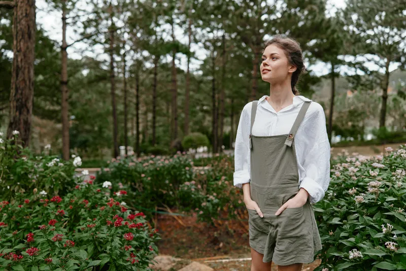 Young woman in casual olive green overalls standing in a blooming garden, expressing tranquility and connection with nature amidst vibrant flowers and greenery