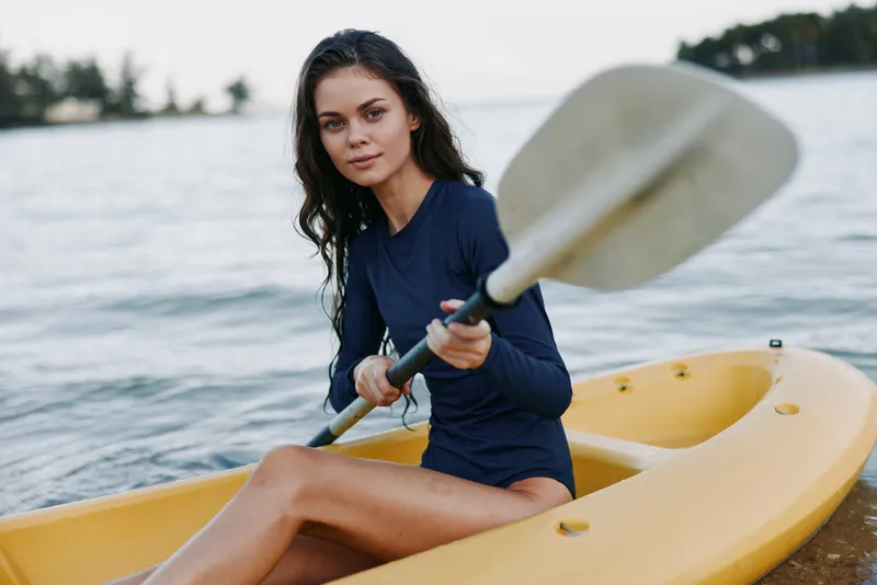 Young woman sits in a yellow kayak on calm water, paddle in hand, wearing a navy swimsuit. Focused expression, soft light, and a relaxed outdoor scene that conveys leisure and adventure.