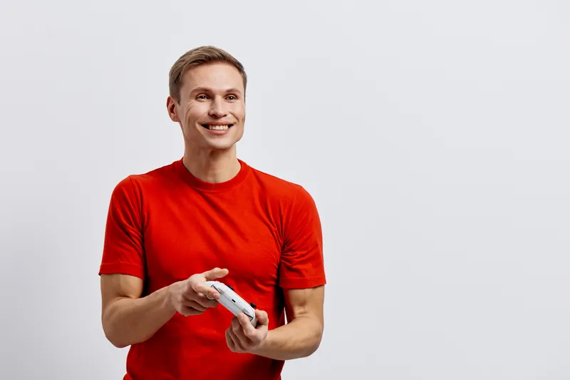 Smiling man wearing red t-shirt holding game controller looking happy and excited isolated on plain light gray background studio portrait. People lifestyle concept