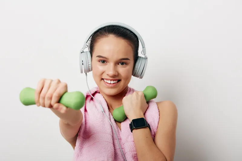 Fitness woman in sport pose, lifting green dumbbells with headphones, pink towel over shoulders, smiling during workout in a clean studio.