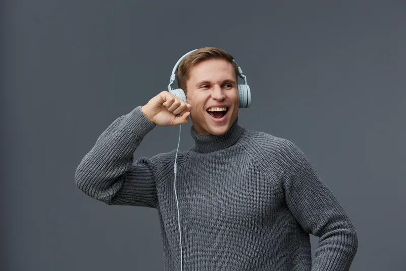 Happy young caucasian man wearing gray sweater and headphones dancing enjoying music isolated on dark grey background winter season mental health concept