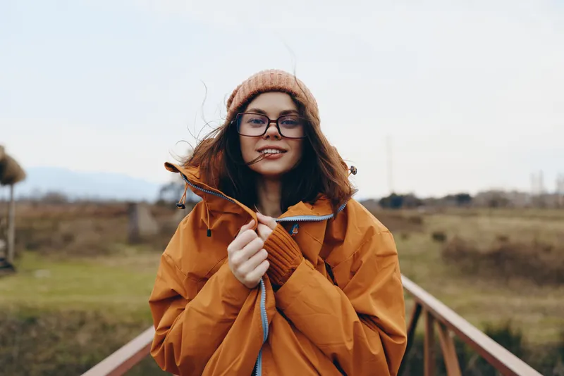 Woman lifestyle outdoors, smiling in autumn coat and hat, glasses on, countryside sunlight, walking along wooden fence, warm daylight, relaxed posture, soft breeze, nature field background