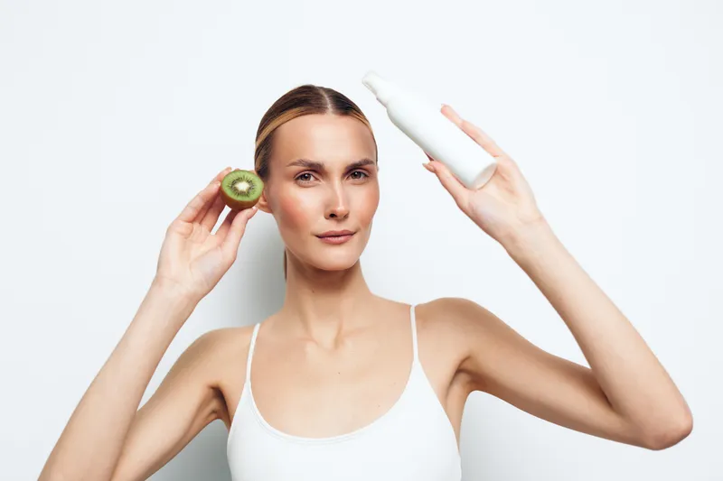 Young woman in white tank top holding kiwi and white cosmetic bottle. Clean background emphasizes natural skincare, beauty, and healthy lifestyle concepts with soft lighting.