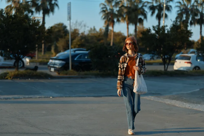 woman walking with smile and sunglasses through sunlit parking lot by palms, casual jeans and tote, candid lifestyle portrait with golden hour glow, authenticity and emotional storytelling.