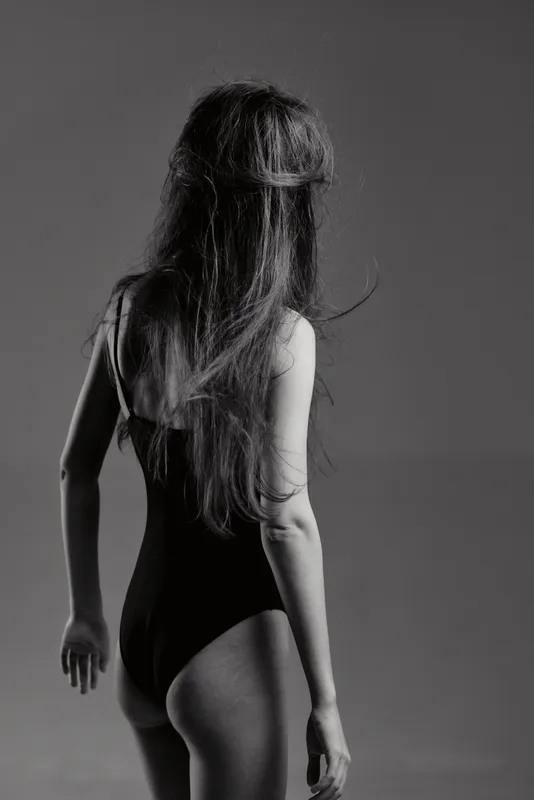 Black and white image of a woman in a swimsuit with long hair, standing with her back to the camera in a minimalistic setting.