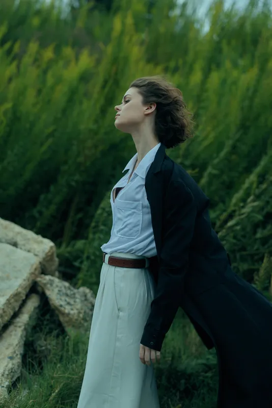 Woman in a white dress and dark coat billowing in the wind, standing among rocks and green reeds for a dramatic fashion outdoors scene.