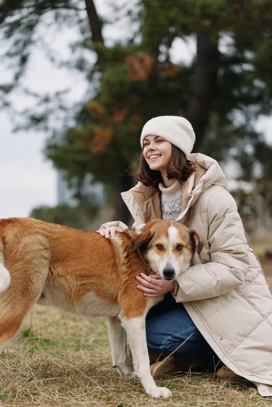 Woman in a warm coat and beanie smiles while kneeling beside a friendly dog in a park on a cold day, capturing a tender moment of companionship