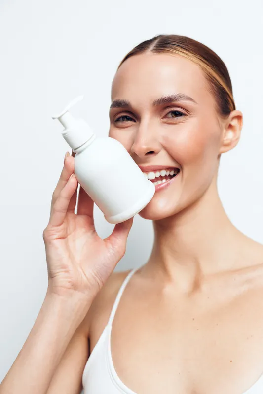 Young woman with clean skin holds white cosmetic bottle with pump smiling showing skincare product for beauty routine isolated on white background.