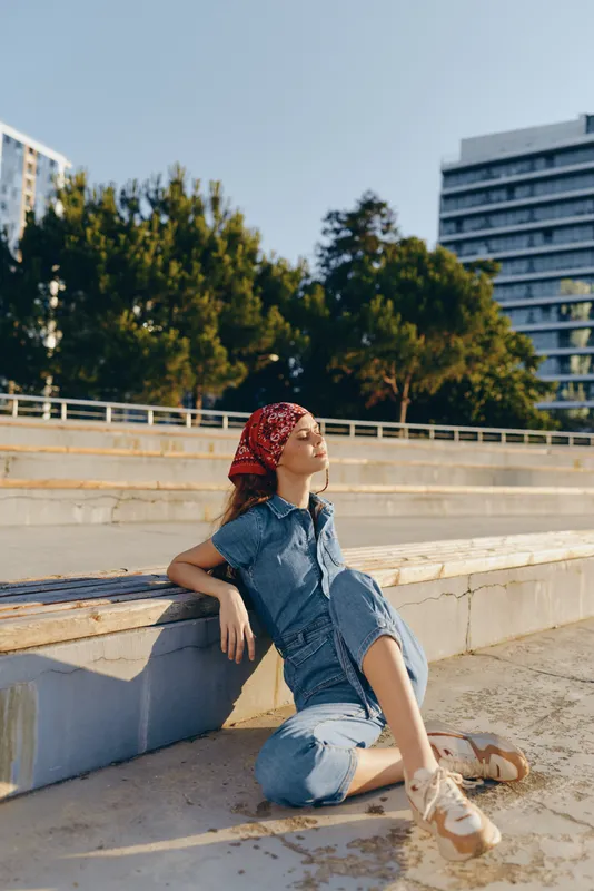 relaxed woman sitting on concrete steps wearing denim jumpsuit and red headscarf enjoying sunny day outdoors with urban buildings and green trees background
