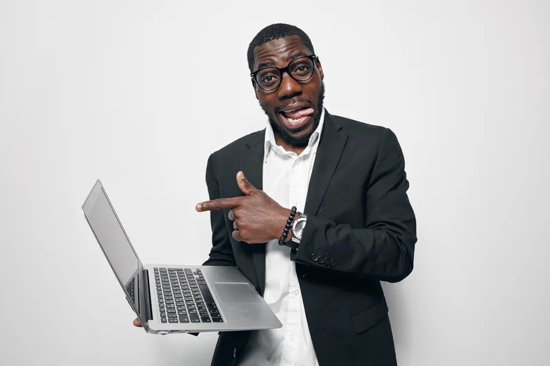 African American man with dark skin wearing glasses and black suit holding laptop and pointing at screen with surprised expression on white background.