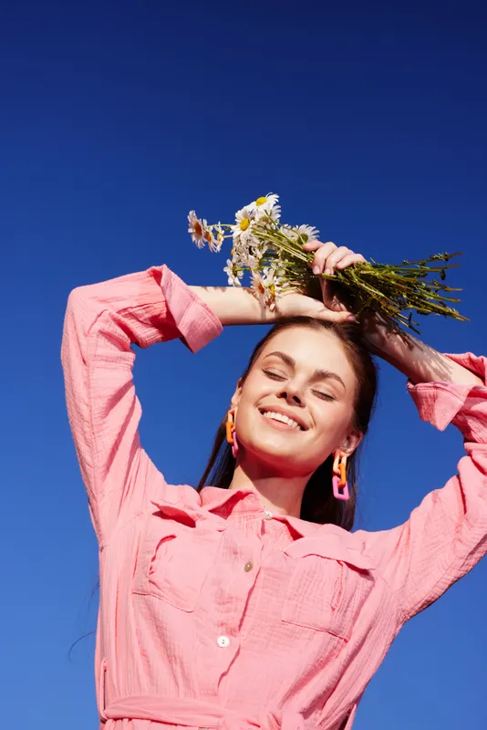 Vibrant young woman with flowers enjoying a sunny day against a blue sky, exuding happiness and joy in a stylish pink outfit