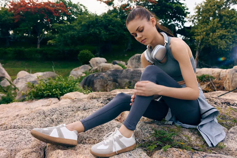 Young woman in athletic wear adjusting her leg while sitting on rocks outdoors, showcasing an active lifestyle and fitness concept