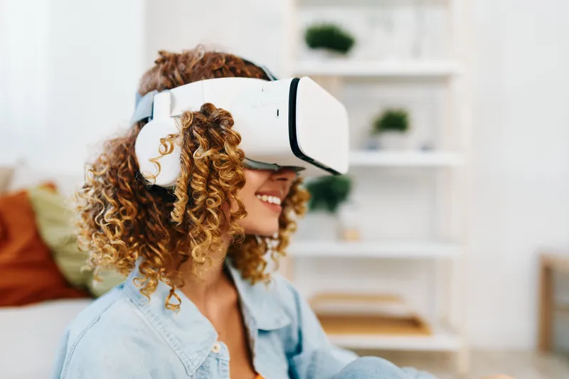 Улыбающаяся женщина наслаждается игрой в виртуальной реальности дома в VR-очках, сидя на диване в футуристической комнате