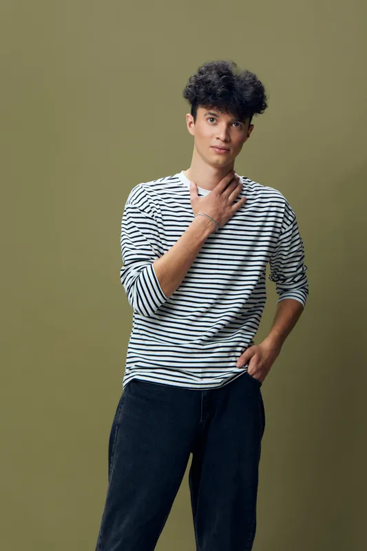 young man standing with hand on neck wearing striped shirt and dark pants against plain olive background with casual style and thoughtful expression