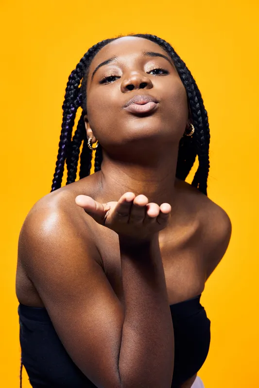 Young African woman blowing a kiss on a bright yellow background, exuding confidence and joy with her stylish braided hairstyle and black sleeveless top