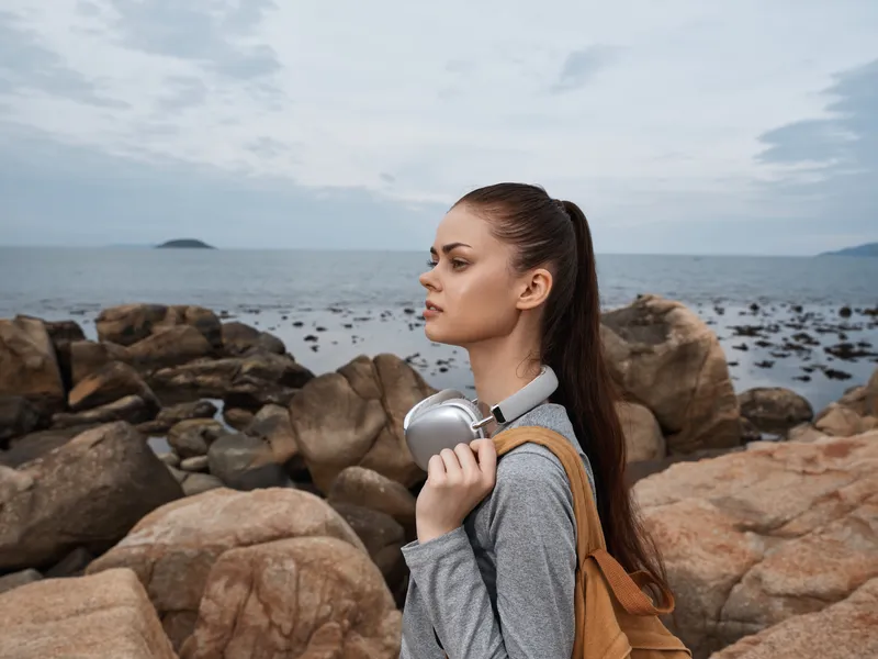 Young woman enjoying nature by the sea with headphones, wearing casual gray clothing and a backpack, emotional and serene atmosphere on rocky shoreline