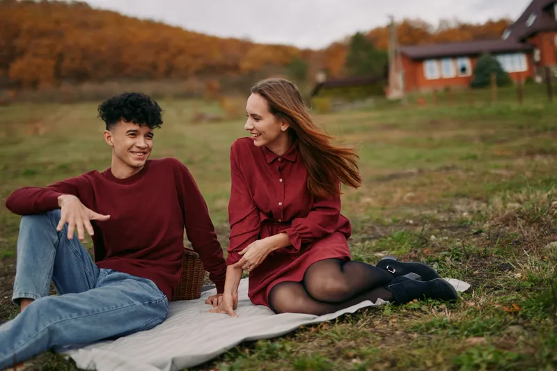 A young couple sharing a genuine outdoor picnic on a blanket near a rustic cottage, candid smiles and authentic interaction captured in warm autumn light, conveying real emotion and close connection.