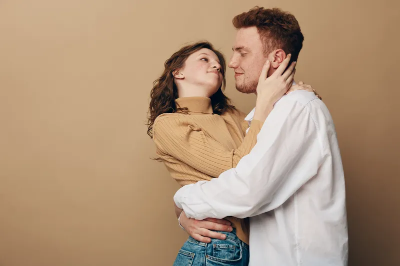 Young couple embraces tenderly, woman in brown sweater and man in white shirt share intimate moment on beige background. Concept of love, affection, and close relationship.