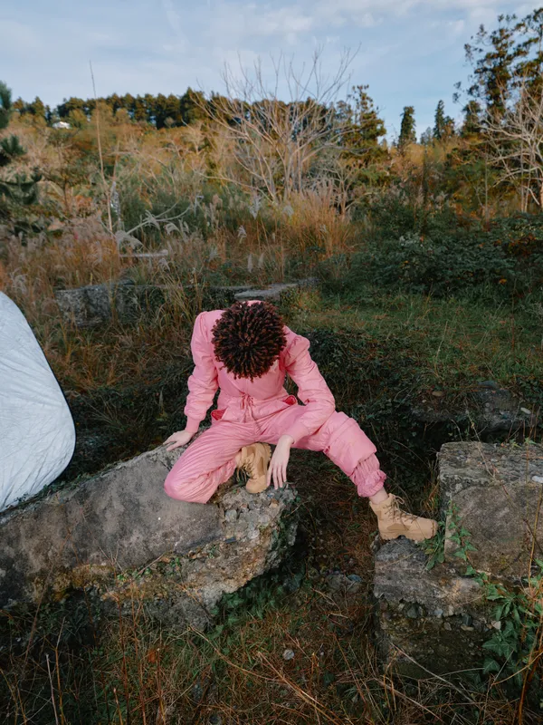 Outdoor scene showing a person in a pink jumpsuit crouching on rocks amid autumn foliage in a rugged natural landscape, conveying exploration, focus, and a quiet outdoor moment.