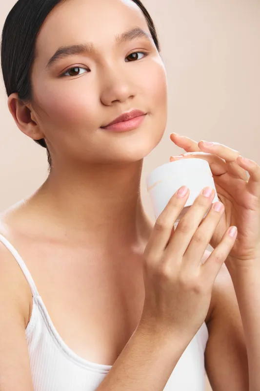 asian woman is seen enjoying a calm skincare moment in a studio. she holds a small white cup near her face, conveying serenity, self care, and gentle beauty in a simple setting.