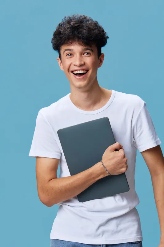 smiling young man in white t-shirt holding laptop with joy and enthusiasm against plain blue background, casual style and positive energy