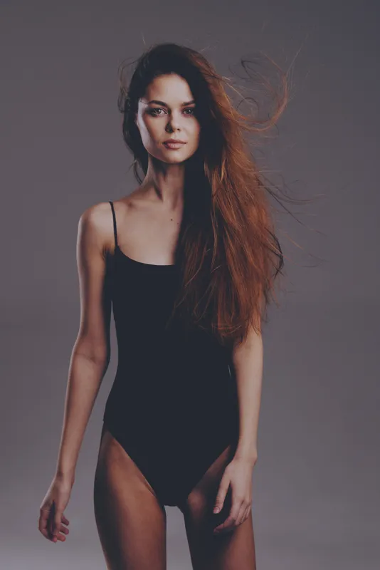 Slim woman with long hair posing in black bodysuit against gray background, showcasing elegance and natural beauty.