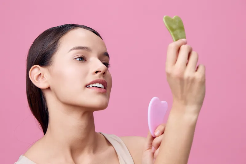 Young woman examining beauty tools on a pink background, showcasing skincare routine and self care emotions with soft tones and heart shaped gua sha stones