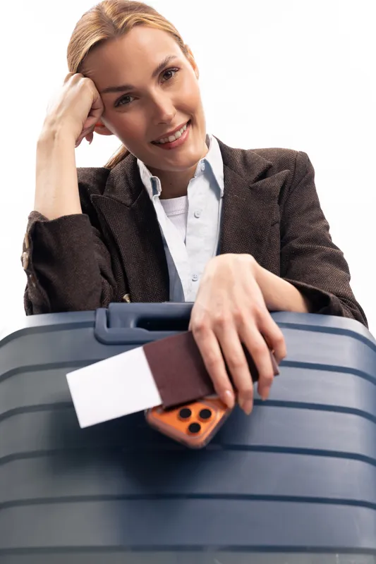 Smiling woman with suitcase holding passport and phone leans on luggage. Travel concept with casual outfit, happy tourist ready for trip isolated on white background.