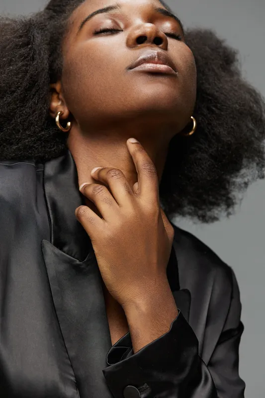 Portrait of a confident young Black woman with natural hair, wearing a black satin blazer, exuding self assurance against a neutral backdrop